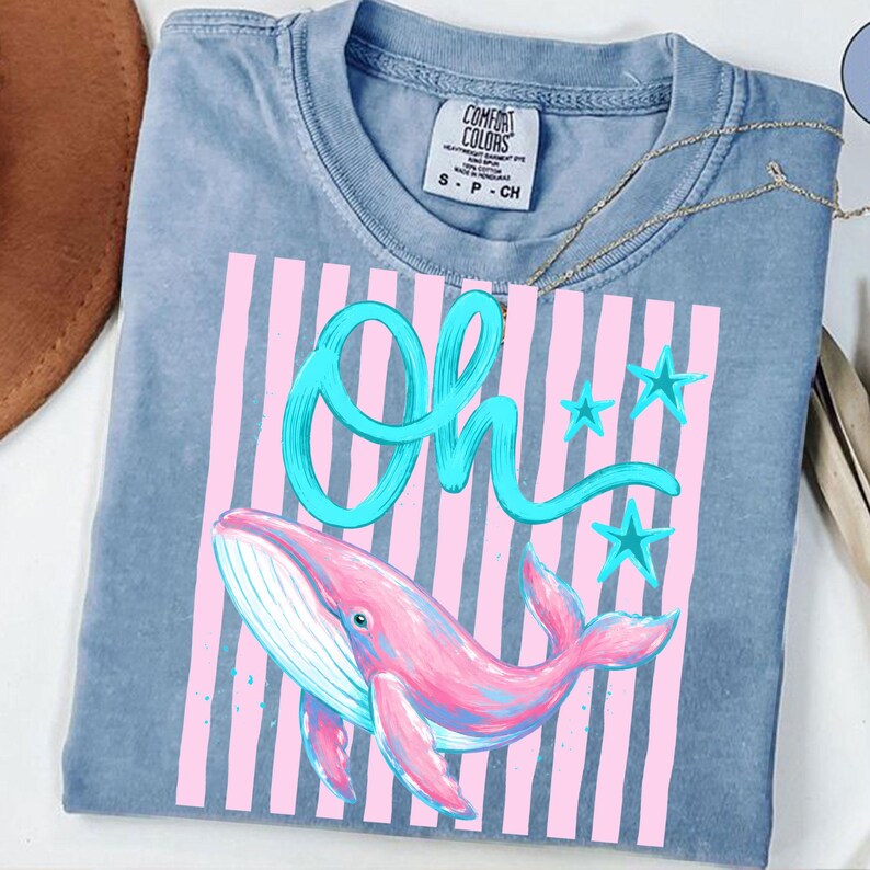 May include: A blue Comfort Colors t-shirt featuring a pink whale illustration with the word "Oh" in turquoise script. The design includes pink vertical stripes and turquoise stars. The shirt has a crew neck and short sleeves.