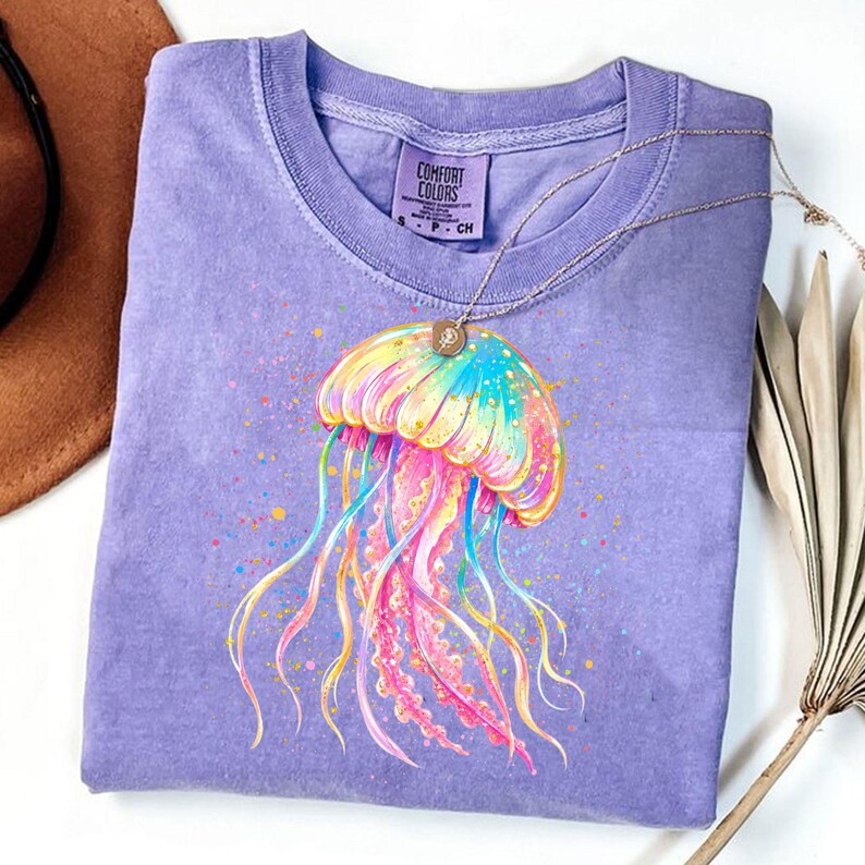 May include: A lilac Comfort Colors t-shirt featuring a colorful jellyfish graphic. The jellyfish design includes shades of pink, yellow, blue, and green. A gold necklace with a small pendant rests on the shirt. The shirt is folded, with a brown hat visible in the background.