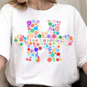 May include: White t-shirt featuring a colorful floral puzzle piece design. The puzzle is filled with various flowers in shades of pink, yellow, blue, and green. The text "IT'S OKAY TO BE DIFFERENT" is written across the puzzle piece.