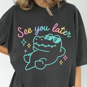 May include: Dark gray t-shirt featuring a cartoon alligator wearing sunglasses and the phrase "See you later" in pastel colors. The alligator is outlined in turquoise, with colorful star accents.