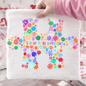May include: A white fabric item featuring a colorful floral puzzle piece design. The puzzle is composed of various flowers in shades of pink, blue, yellow, and orange. The text "IT'S OKAY TO BE DIFFERENT" is printed across the center in rainbow colors.