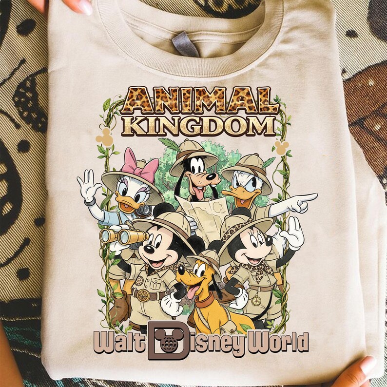 May include: Beige t-shirt featuring a graphic of Disney characters in safari attire. The design includes Mickey Mouse, Donald Duck, Goofy, Pluto, and Daisy Duck, with the words "ANIMAL KINGDOM" and "Walt Disney World".