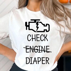 May include: White t-shirt with a black graphic of a car engine warning light and the text "Check Engine" crossed out and "Diaper" written below.