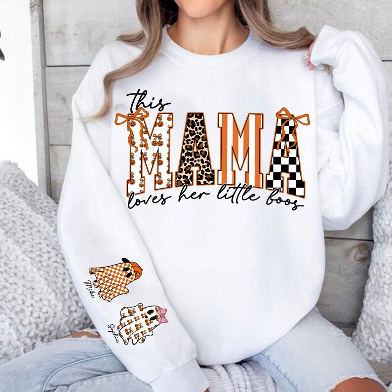 May include: White sweatshirt with the text "This MAMA loves her little boos." The word "MAMA" is in orange, leopard print, and black and white checkered patterns. Two ghost graphics are on the sleeve, one with the name "Mike" and the other with the name "Sophia".