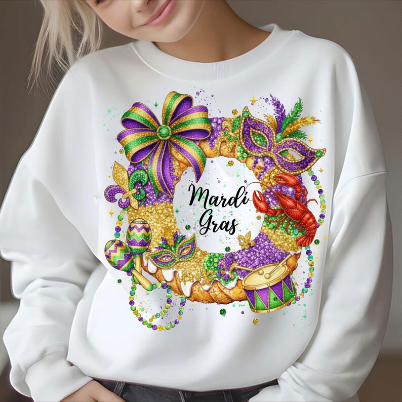 May include: White sweatshirt featuring a colorful Mardi Gras design. The design includes a wreath with a bow, mask, maracas, and a crawfish. The words "Mardi Gras" are written in the center of the wreath. The design incorporates purple, green, gold, and red.