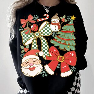 May include: Black sweatshirt featuring a festive Christmas design. The graphic includes a smiling Santa Claus, a snowman, a Christmas tree, and several red bows with green and white checkered accents. Snowflakes and candy canes complete the holiday theme.