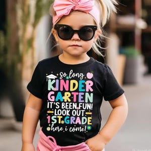 Puede incluir: Una camiseta negra con las palabras "So long Kindergarten, It's been fun! Look out 1st Grade here I come!" en letras coloridas. La camiseta tiene una gorra de graduaci&oacute;n, una manzana y gr&aacute;ficos de flores. El ni&ntilde;o lleva un lazo rosa y gafas de sol.