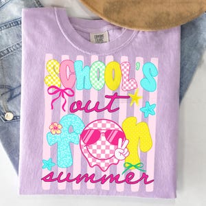 May include: A lilac Comfort Colors t-shirt with a colorful graphic that says "School's Out for Summer." The design includes checkered patterns, stars, bows, and a smiley face wearing sunglasses.