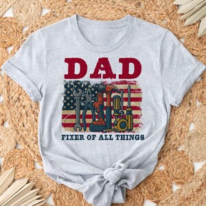 May include: Gray t-shirt with the text "DAD FIXER OF ALL THINGS" in red and black. An American flag with tools is behind the text.