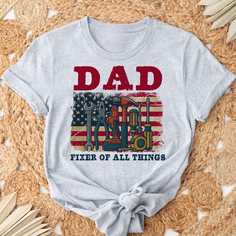 May include: Gray t-shirt with the text "DAD FIXER OF ALL THINGS" in red and black. An American flag with tools is behind the text.