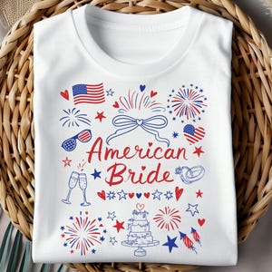 May include: White t-shirt with a patriotic design featuring the text "American Bride" in red script. The design includes an American flag, fireworks, hearts, stars, champagne glasses, and a wedding cake, all in red and blue.