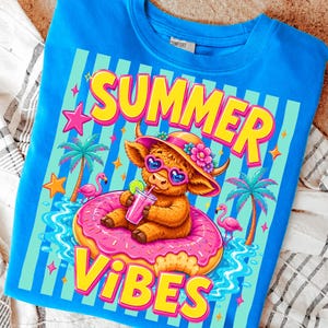 May include: A blue t-shirt with a colorful graphic. The graphic features a cartoon Highland cow wearing sunglasses and a hat, floating on a pink donut in water. The words "SUMMER VIBES" are written in yellow and pink.