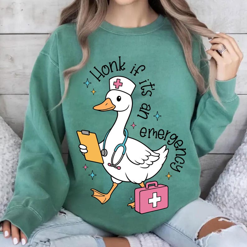 Puede incluir: Sudadera verde con un ganso de dibujos animados que lleva un gorro de enfermera y sostiene un portapapeles y un estetoscopio. El texto "Honk if it's an emergency" est&aacute; impreso en la sudadera.