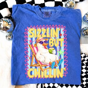 May include: A blue Comfort Colors t-shirt featuring a cartoon duck wearing sunglasses and a hat, relaxing in a beach chair with a coconut drink. The shirt has the text "Sizzlin' But Chillin'" in yellow and pink.