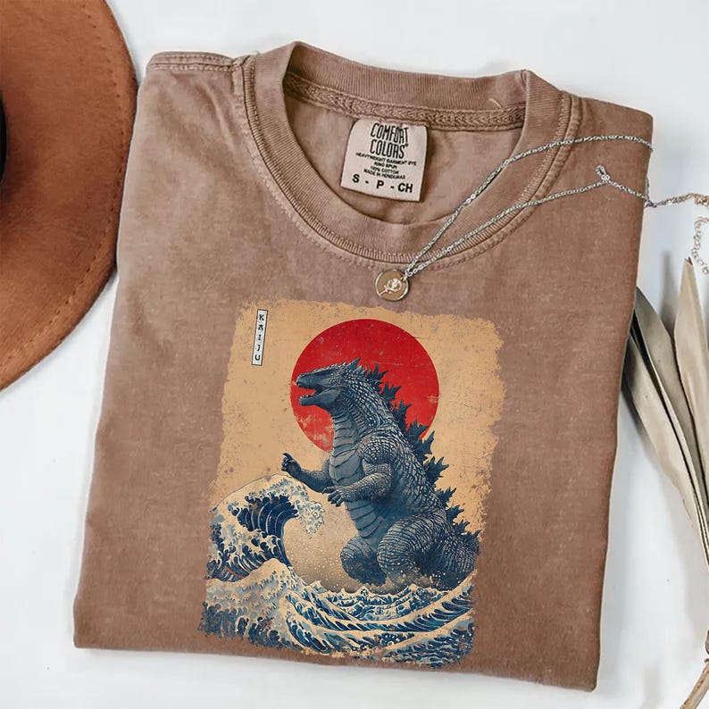 May include: Brown Comfort Colors t-shirt with a Godzilla graphic in a Japanese art style. The design includes a red sun, ocean waves, and the word "Kaiju". A silver necklace with a pendant is draped across the shirt.