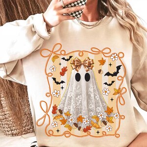 May include: Beige sweatshirt featuring a Halloween ghost design. The ghost has a gold bow, black eyes, and a lace-like veil. The design includes bats, leaves, flowers, and acorns within an orange frame.