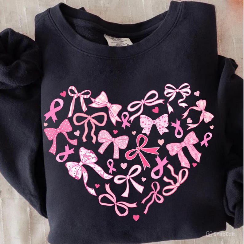 May include: A navy blue sweatshirt featuring a heart-shaped design composed of pink bows and ribbons. The bows vary in style and pattern, with some including polka dots. Small pink hearts are scattered throughout the design.