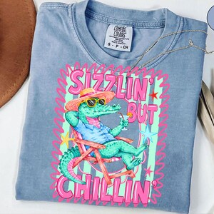 May include: A light blue Comfort Colors t-shirt featuring a cartoon alligator wearing sunglasses and a hat, relaxing in a beach chair while holding a cocktail. The shirt has the words "Sizzlin' But Chillin'" in pink and green.