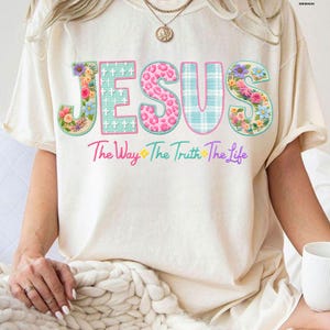 Puede incluir: Camiseta color crema con la palabra "JESUS" en un dise&ntilde;o floral y estampado. El texto "The Way The Truth The Life" est&aacute; debajo de la palabra "JESUS". La camiseta tiene mangas cortas y un corte holgado.