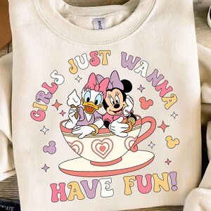 May include: A white sweatshirt with a pink, yellow, and blue graphic print of two cartoon characters, a duck and a mouse, sitting in a teacup. The text "Girls Just Wanna Have Fun!" is printed in a retro font around the characters.