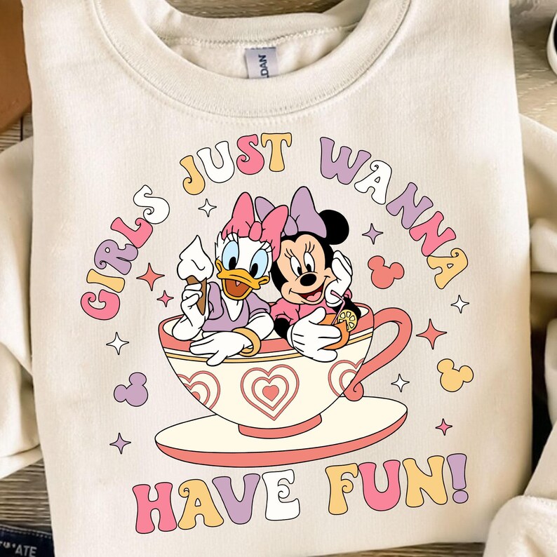 May include: A white sweatshirt with a pink, yellow, and blue graphic print of two cartoon characters, a duck and a mouse, sitting in a teacup. The text "Girls Just Wanna Have Fun!" is printed in a retro font around the characters.