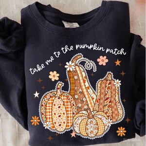 以下が含まれることがあります： 秋をテーマにしたデザインのネイビーブルーのスウェットシャツ。グラフィックには、花柄やチェック柄のパンプキンがいくつかあり、「take me to the pumpkin patch」というテキストが添えられています。装飾的な花と星がデザインを完成させています。