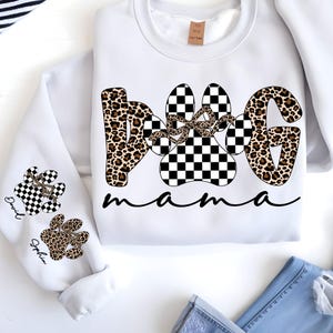 May include: White sweatshirt with "DOG mama" text in black script. The word "DOG" is designed with a leopard print and a checkered paw print. The sleeves feature paw prints with names.
