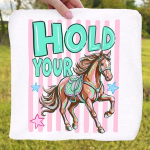 May include: White square towel with a cartoon horse illustration. The horse is brown with a teal saddle and is set against a background of pink stripes. The text "HOLD YOUR" is in teal, with star accents.
