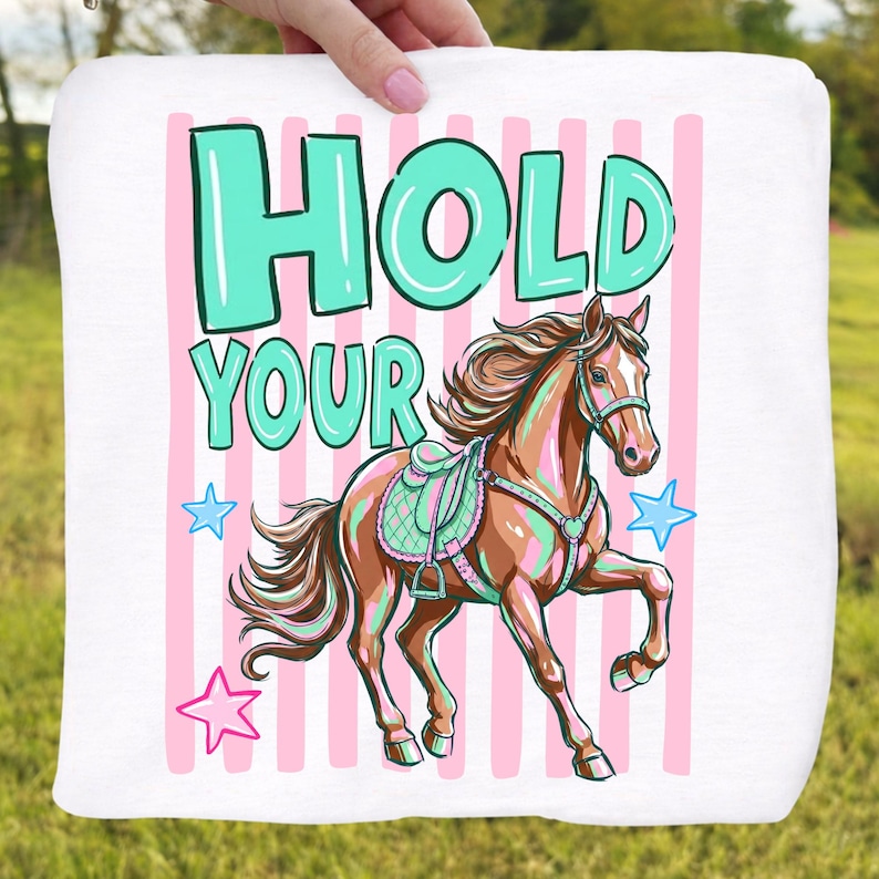 May include: White square towel with a cartoon horse illustration. The horse is brown with a teal saddle and is set against a background of pink stripes. The text "HOLD YOUR" is in teal, with star accents.