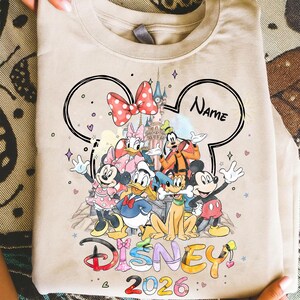May include: Beige crew neck sweatshirt featuring a colorful graphic of Disney characters, including Mickey Mouse, Minnie Mouse, Donald Duck, and Goofy. The design includes the word "Disney" and the year "2026". A black outline of Mickey Mouse ears frames the design.
