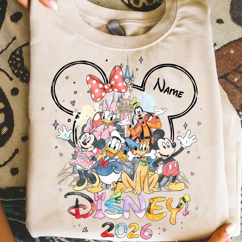 May include: Beige crew neck sweatshirt featuring a colorful graphic of Disney characters, including Mickey Mouse, Minnie Mouse, Donald Duck, and Goofy. The design includes the word "Disney" and the year "2026". A black outline of Mickey Mouse ears frames the design.