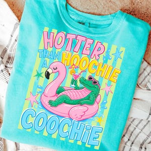 May include: A turquoise crew neck sweatshirt with a graphic design. The design features a pink flamingo float with a green alligator wearing sunglasses, holding a cocktail. Text reads "Hotter Than A Hoochie Coochie."