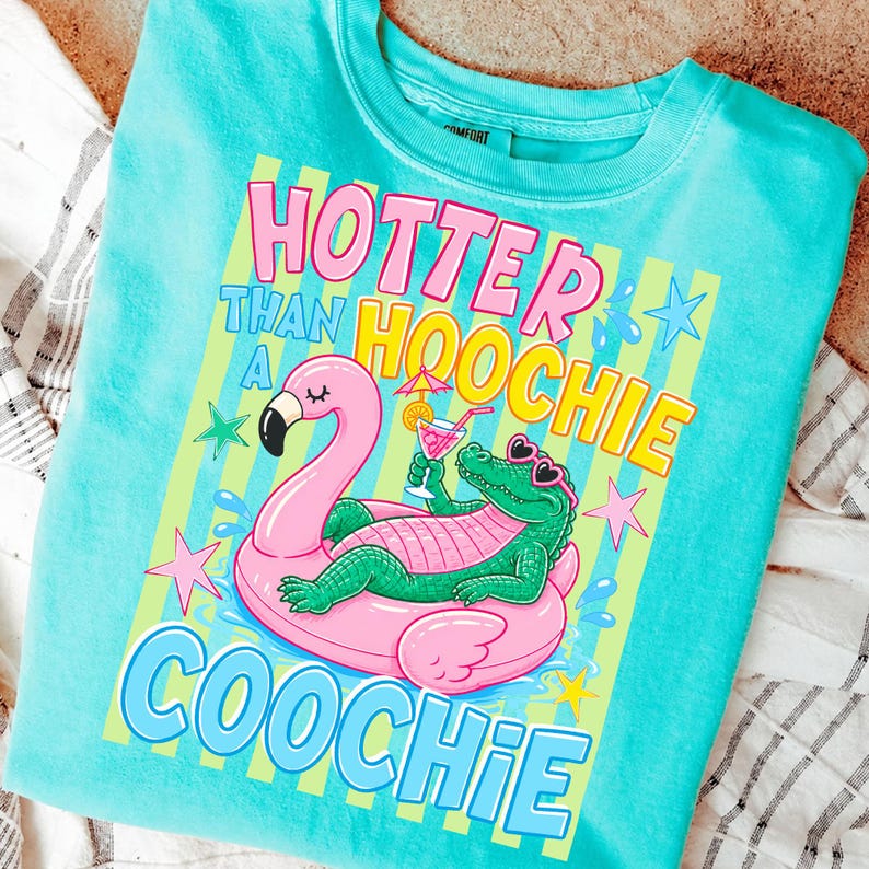 May include: A turquoise crew neck sweatshirt with a graphic design. The design features a pink flamingo float with a green alligator wearing sunglasses, holding a cocktail. Text reads "Hotter Than A Hoochie Coochie."