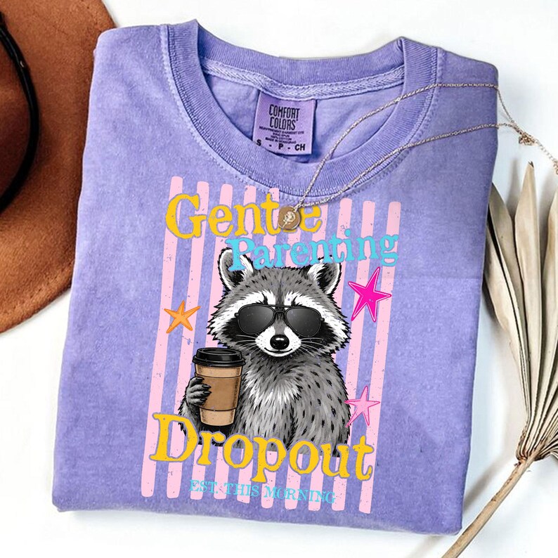 Op de afbeelding: Een lichtpaars Comfort Colors T-shirt met een wasbeer met een zonnebril en een koffiekopje. Het shirt heeft de tekst "Gentle Parenting Dropout" met sterren en verticale strepen.