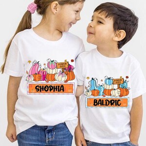 May include: Two white t-shirts with a colorful pumpkin patch design. One shirt says "Shophia" and the other says "Baldric" in an orange banner. The shirts feature pumpkins in various colors.