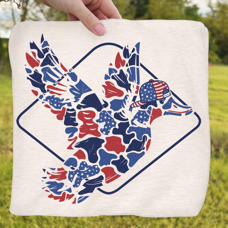 May include: A cream-colored square fabric featuring a patriotic duck design. The duck is composed of red, white, and blue shapes, including stars and stripes, and wears a baseball cap. The design is framed by a dark blue diamond.