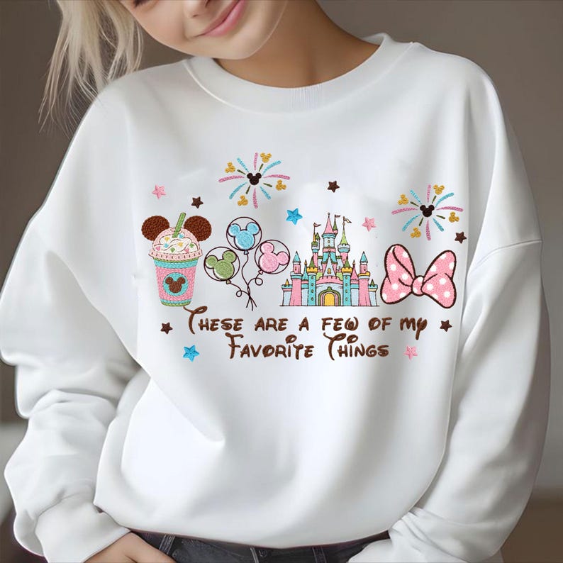 May include: White sweatshirt with embroidered Disney-themed graphics. Features a drink with Mickey Mouse ears, balloons, a castle, and a bow. Text reads "These are a few of my favorite things."