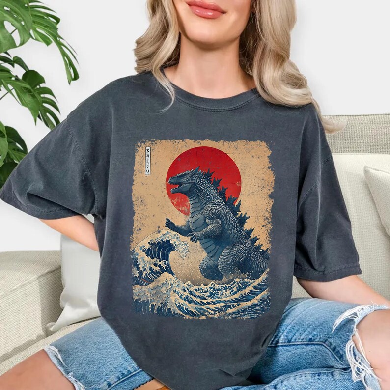 May include: Dark grey t-shirt with a Japanese-inspired design of a Godzilla-like creature, a red sun, and ocean waves. The word "KAIJU" is written in Japanese. The shirt has a loose fit and is made of a soft material.