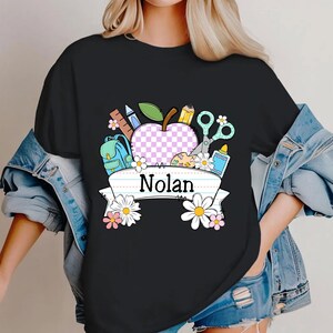 May include: Black t-shirt with a colorful school-themed graphic. The design includes an apple with a checkered pattern, school supplies, and the name "Nolan" on a banner, surrounded by flowers.