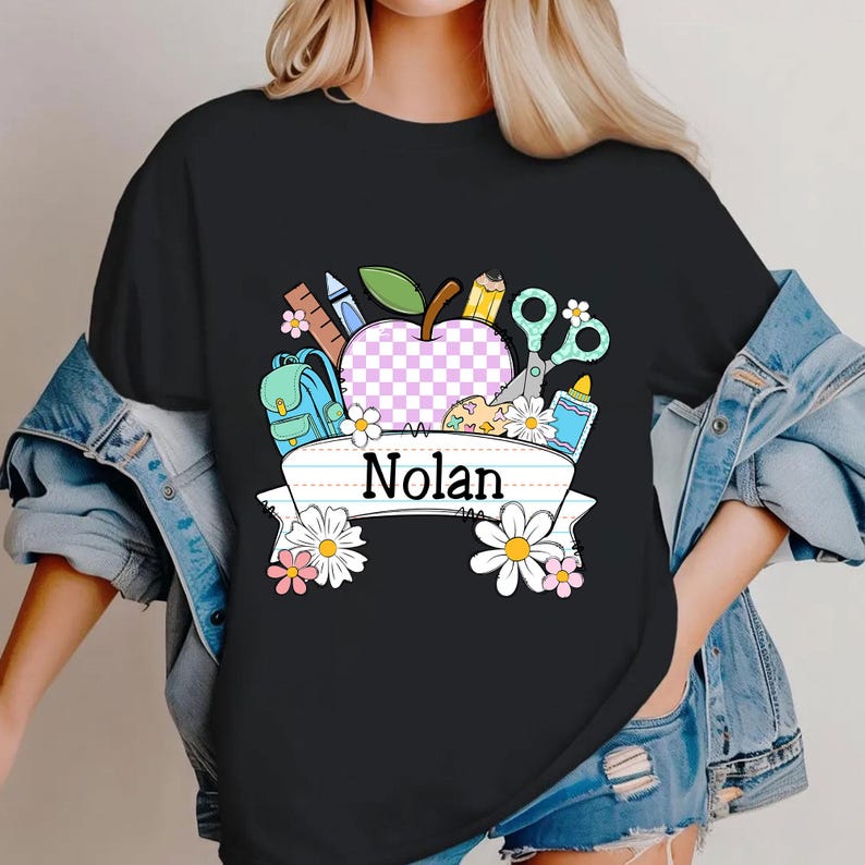 May include: Black t-shirt with a colorful school-themed graphic. The design includes an apple with a checkered pattern, school supplies, and the name "Nolan" on a banner, surrounded by flowers.
