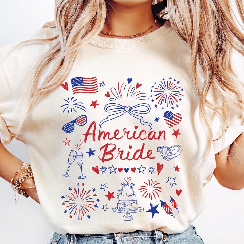 May include: Cream-colored sweatshirt with a patriotic design. Features the text "American Bride" in red and blue, along with illustrations of the American flag, fireworks, hearts, stars, and a wedding cake. A festive design for a wedding.