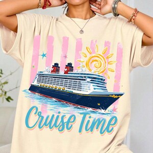 May include: Cream-coloured t-shirt featuring a cruise ship illustration with a sun and the text "Cruise Time" in blue. The ship is navy blue with red accents, set against pink vertical stripes and a blue ocean.