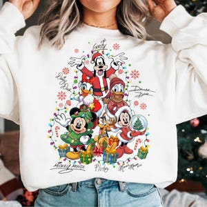 Puede incluir: Sudadera blanca con un diseño de árbol de Navidad con personajes de Disney. Mickey, Minnie, Goofy, Donald, Daisy y Pluto están rodeados de luces navideñas, copos de nieve y bastones de caramelo. Los personajes llevan trajes festivos.
