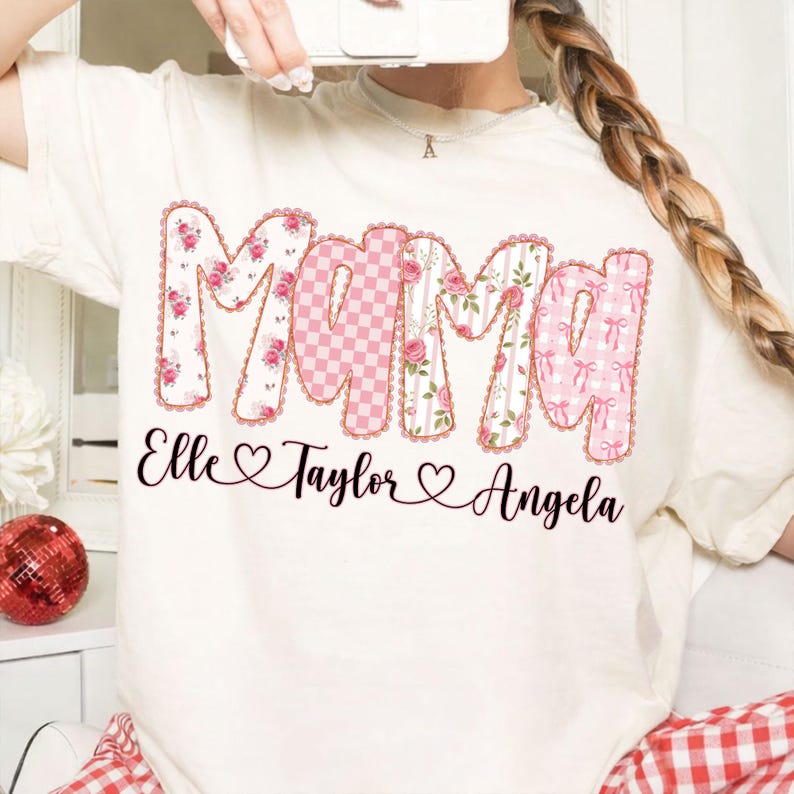 May include: Cream-colored t-shirt with the word "Mama" in large, decorative letters. The letters are patterned with floral, gingham, and striped designs in pink and white. Below the word are the names "Elle", "Taylor", and "Angela".