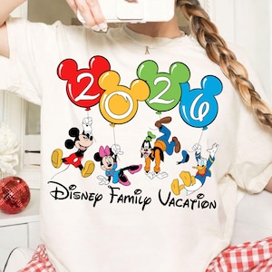 Może przedstawiać: Kremowa koszulka z napisem "Disney Family Vacation" i postaciami Disneya: Myszka Miki, Myszka Minnie, Goofy i Kaczor Donald. Postacie trzymają balony w kształcie cyfr 2, 0, 2 i 0 w kolorach czerwonym, ż&oacute;łtym, zielonym i niebieskim.