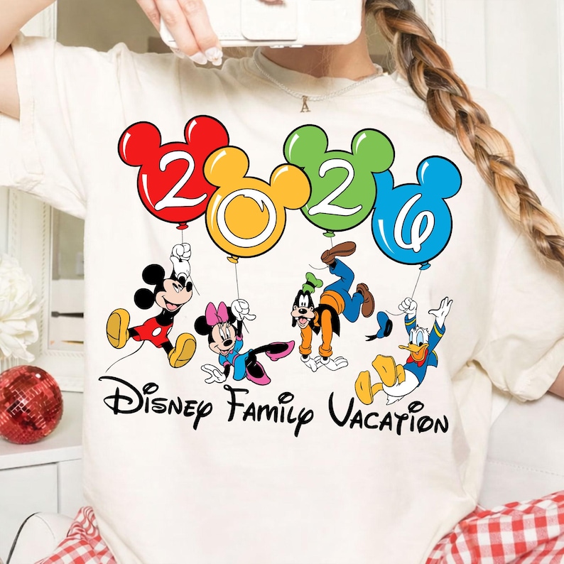 Może przedstawiać: Kremowa koszulka z napisem "Disney Family Vacation" i postaciami Disneya: Myszka Miki, Myszka Minnie, Goofy i Kaczor Donald. Postacie trzymają balony w kształcie cyfr 2, 0, 2 i 0 w kolorach czerwonym, ż&oacute;łtym, zielonym i niebieskim.