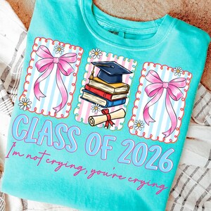 May include: Turquoise crew neck t-shirt with a graduation theme. The shirt features three framed graphics with pink bows, a graduation cap, books, and a diploma. Text on the shirt reads "CLASS OF 2026" and "I'm not crying, you're crying."