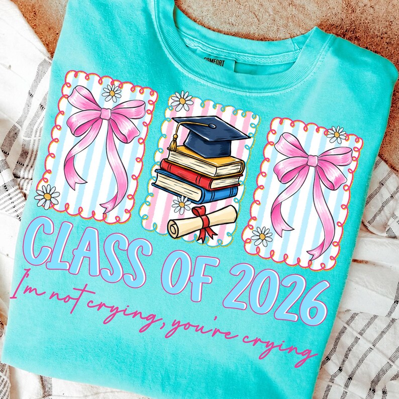 May include: Turquoise crew neck t-shirt with a graduation theme. The shirt features three framed graphics with pink bows, a graduation cap, books, and a diploma. Text on the shirt reads "CLASS OF 2026" and "I'm not crying, you're crying."