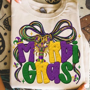 May include: A folded, cream-colored t-shirt featuring a colorful Mardi Gras design. The design includes the words "Mardi Gras" in purple, green, and yellow, with a decorative bow and a detailed crawfish illustration.