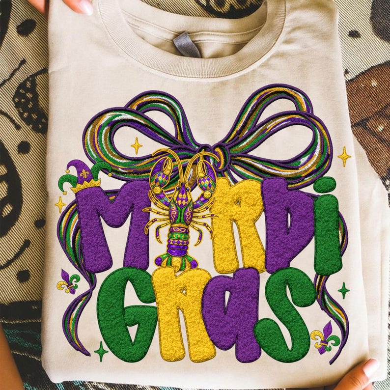 May include: A folded, cream-colored t-shirt featuring a colorful Mardi Gras design. The design includes the words "Mardi Gras" in purple, green, and yellow, with a decorative bow and a detailed crawfish illustration.